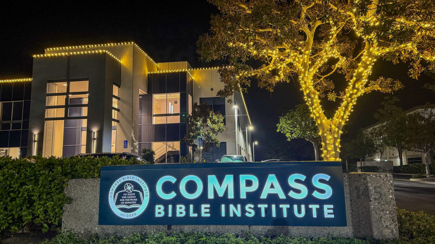 Compass Bible Institute sign in front of a building with a lit roofline and a large tree wrapped in warm white lights