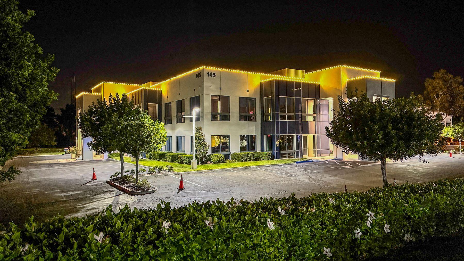Compass Bible Institute at night, with warm white lights along the building roofline and wrapped trees in the parking lot