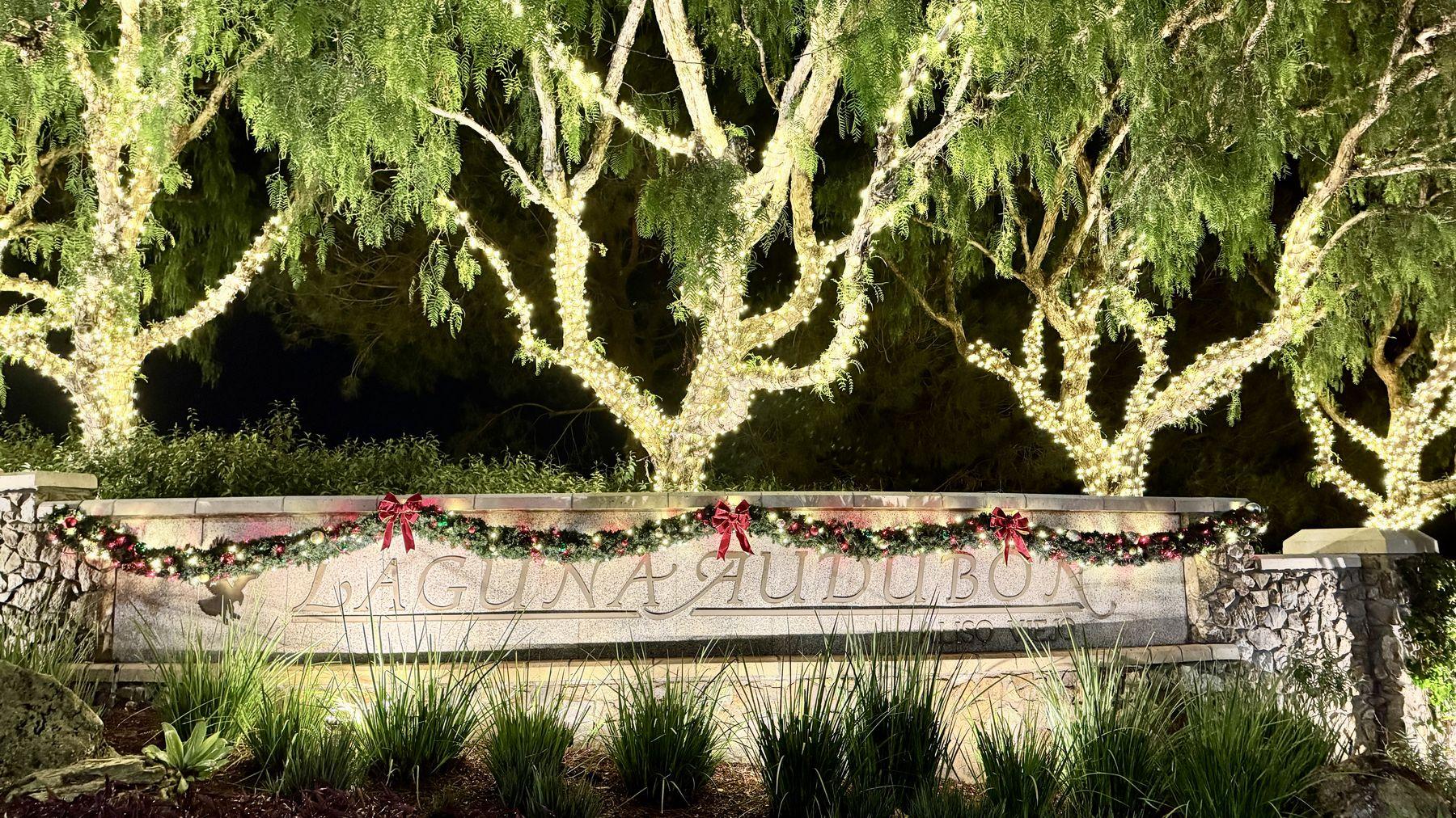 Laguna Audubon community entrance at night, with pepper trees wrapped in warm white lights above a stone sign hung with garland and red bows